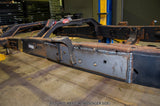 A heavy-duty welded steel frame, likely a vehicle chassis or trailer frame, shown in a workshop setting. The frame features a dark gray painted section on the passenger side with visible welds and mounting holes, and a rusted black section on the driver side.