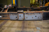 A heavy-duty welded steel frame section, likely a vehicle chassis or frame sub-assembly, shown in a workshop setting. The frame features a large silver-gray rectangular plate on the passenger side with pre-drilled mounting holes and a welded upright bracket.