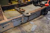 A close-up of a vehicle frame or chassis beam in a workshop, showing welded metal sections, mounting holes, and rusted areas on a concrete floor.